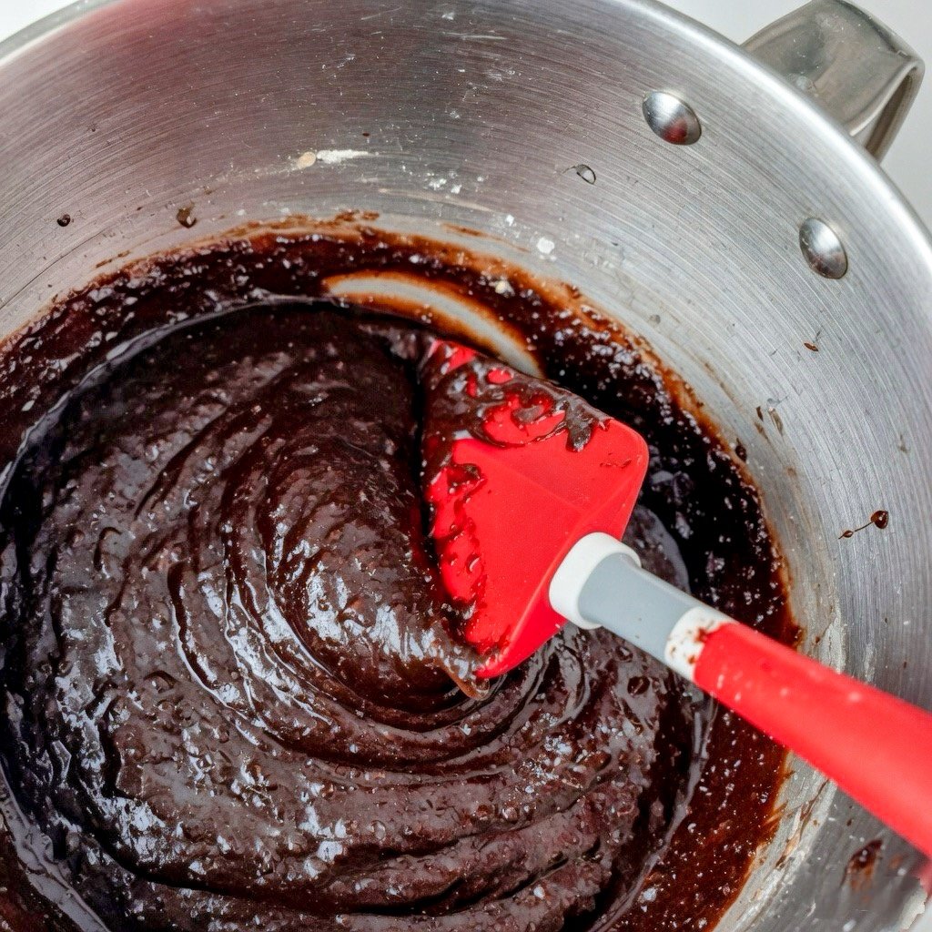 why is my cake dense? Thick glossy chocolate cake batter in a metal bowl being mixed with a red spatula, showing overmixed dense texture