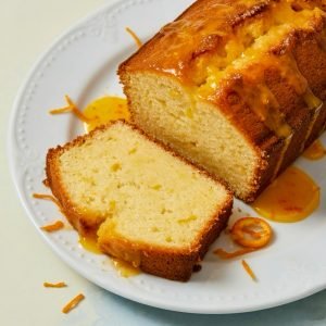 Close-up of a moist orange pound cake loaf on a white plate, topped with glossy citrus glaze and orange zest, with one slice cut and placed in front showing a soft, tender crumb.
